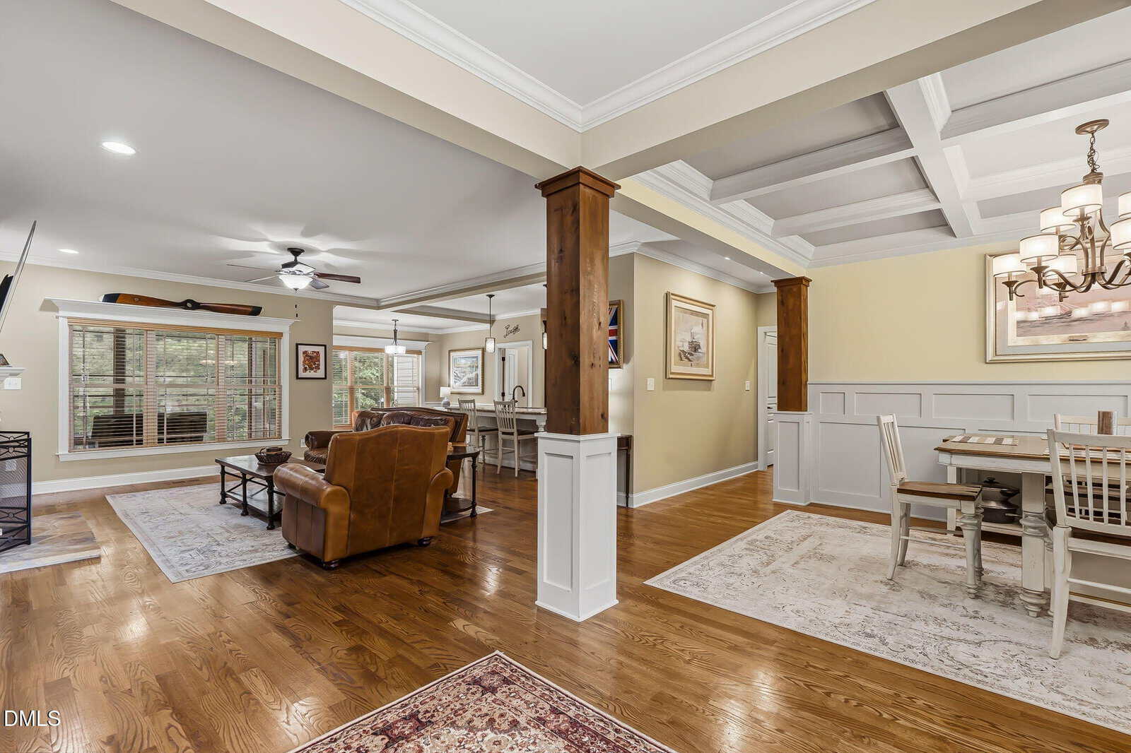 Open-concept living and dining area with hardwood floors, coffered ceiling, neutral walls, and large windows. The space is furnished with a leather armchair, sofas, area rugs, and a dining table with chairs.