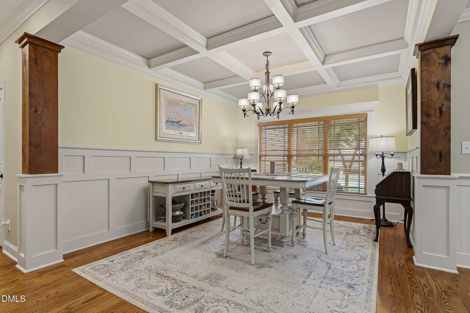 A bright, elegant dining room with coffered ceiling, chandelier, wooden floor, wainscoting walls, a large window with blinds, a white dining table with chairs, and a rug underneath.