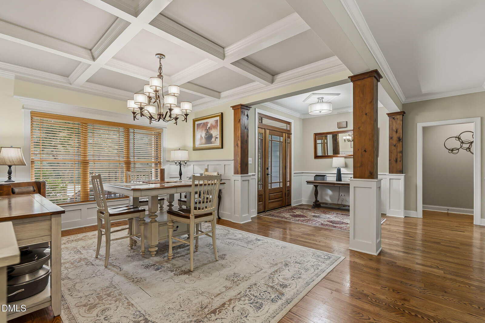 Elegant dining room with coffered ceiling, chandelier, large windows with blinds, a rectangular dining table with chairs, hardwood floors, and an entryway with wood and glass double doors. Neutral tones and classic decor.