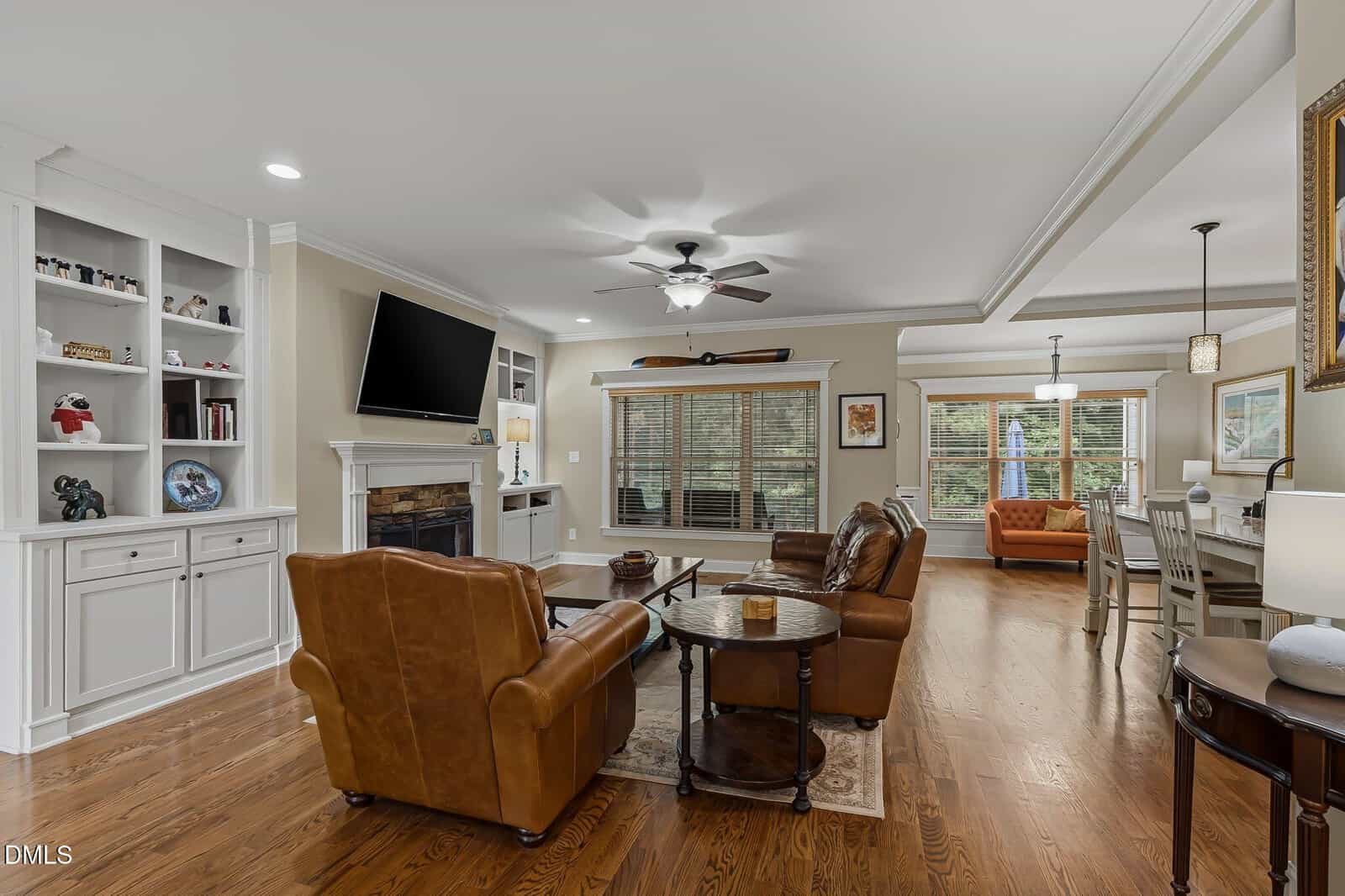 Spacious living room with leather armchairs, a fireplace, built-in shelves, and a large TV. Hardwood floors, large windows with blinds, and an open kitchen and dining area with pendant lighting are visible.