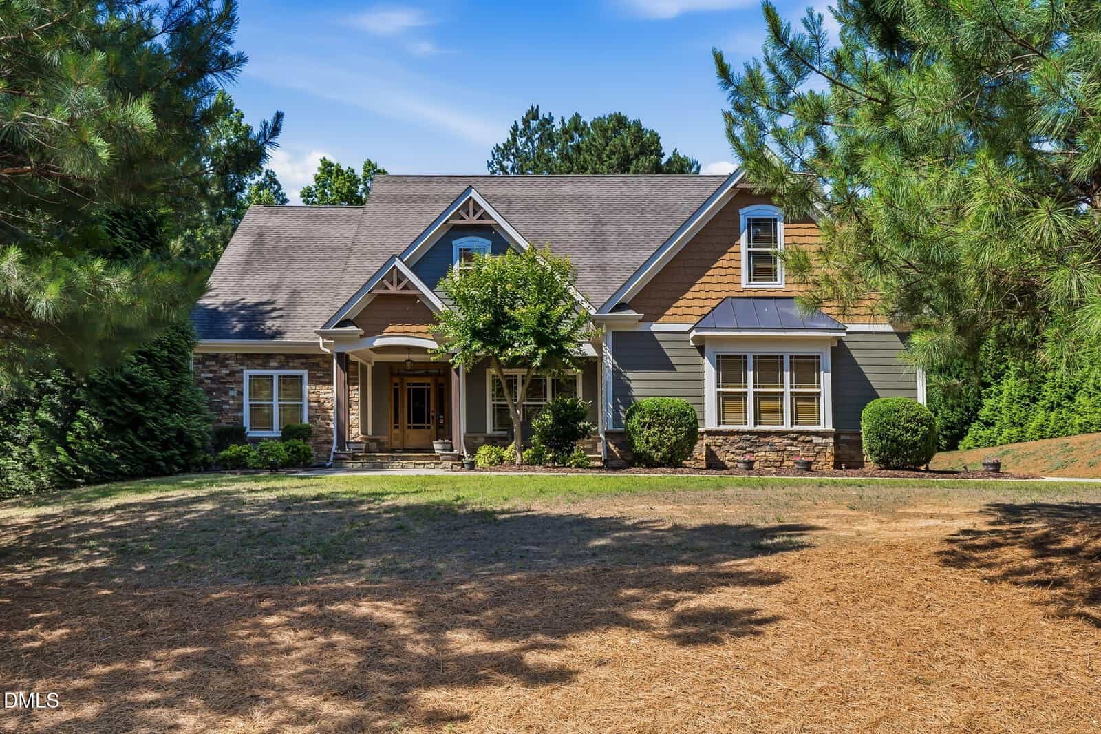 A two-story suburban house with a gabled roof, stone and wood siding, a covered front porch, and neatly landscaped shrubs, surrounded by pine trees and a dry grass lawn.