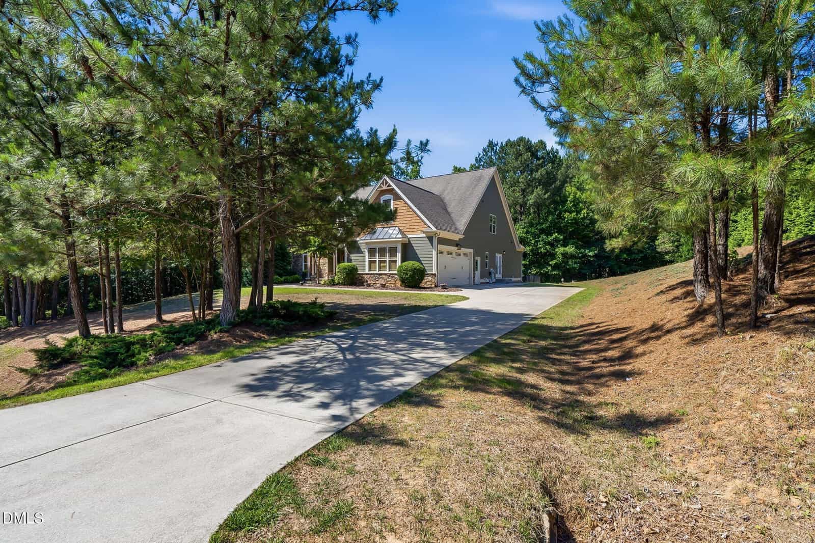 A tan house with a steep roof sits at the end of a long concrete driveway, surrounded by tall pine trees and landscaped greenery under a clear blue sky.