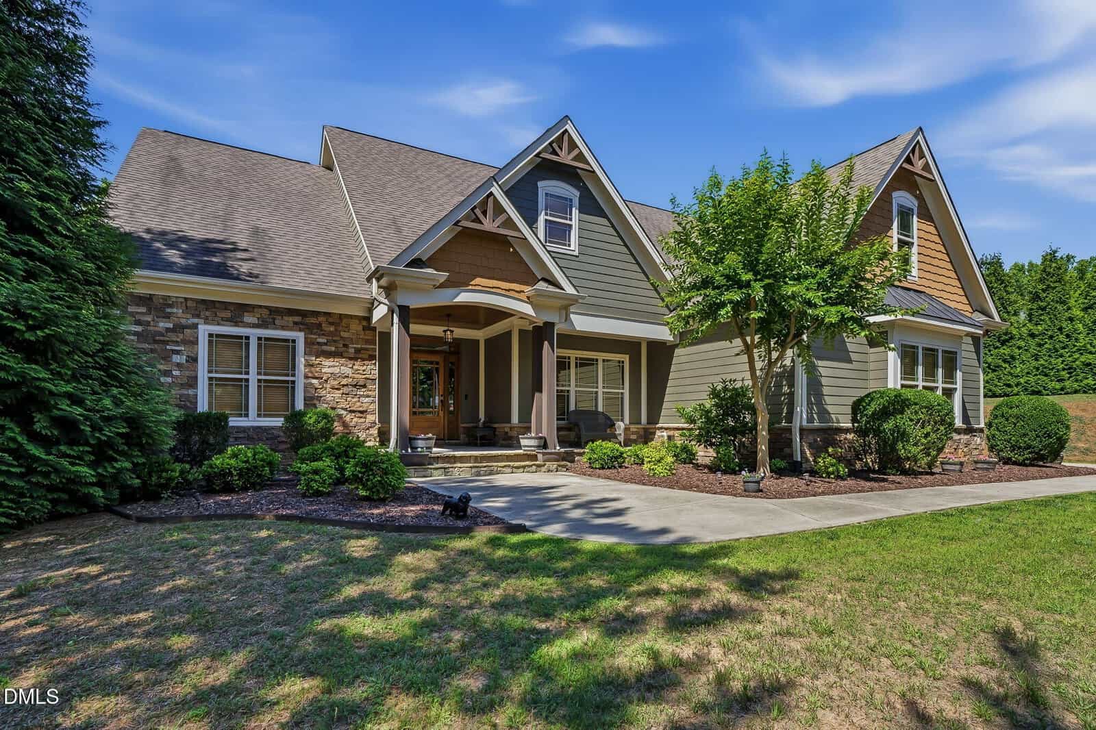 A single-story suburban house with a covered porch, gabled roof, stone and wood siding, a small front patio, neatly trimmed bushes, and a concrete walkway, surrounded by green lawn and trees under a blue sky.