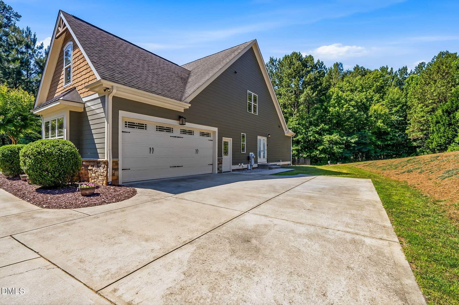 Gray two-story house with a double garage, large driveway, and green lawn bordered by trees under a blue sky.