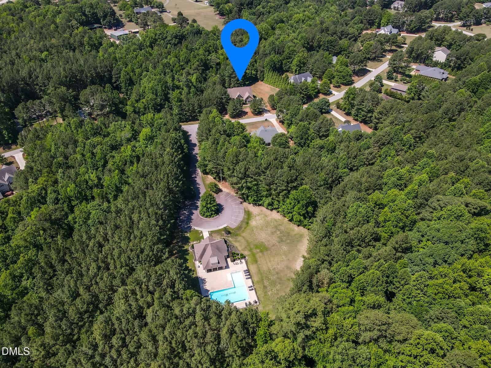 Aerial view of a tree-filled suburban neighborhood with houses. A large blue location marker highlights one property. A swimming pool and building are visible in a clearing among the trees.
