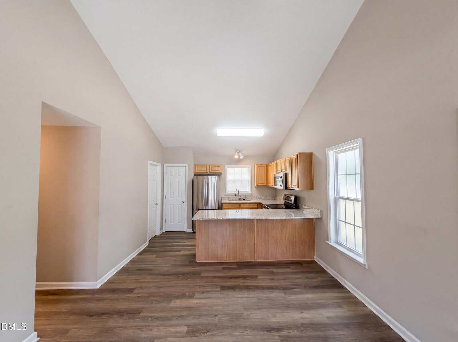 Open kitchen with light wood cabinets, marble countertops, and stainless steel appliances. The space has tan walls, wood flooring, and large windows allowing natural light. A door and pantry are visible in the background.