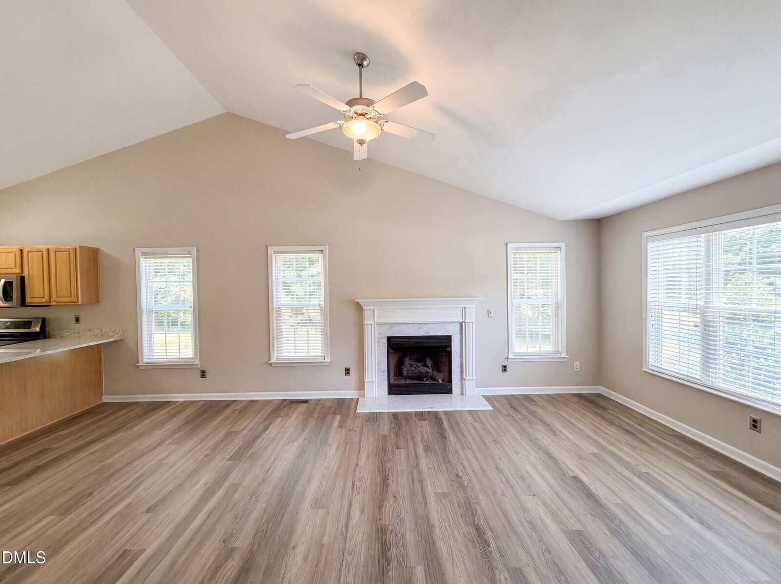 A bright, empty living room with light wood flooring, a white ceiling fan, three tall windows, a white fireplace, and a view of the kitchen with light wood cabinets on the left.