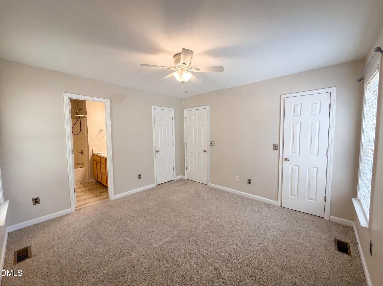 Bright, carpeted bedroom with beige walls, ceiling fan, three white doors, two windows with blinds, and an en suite bathroom visible with a sink and shower.