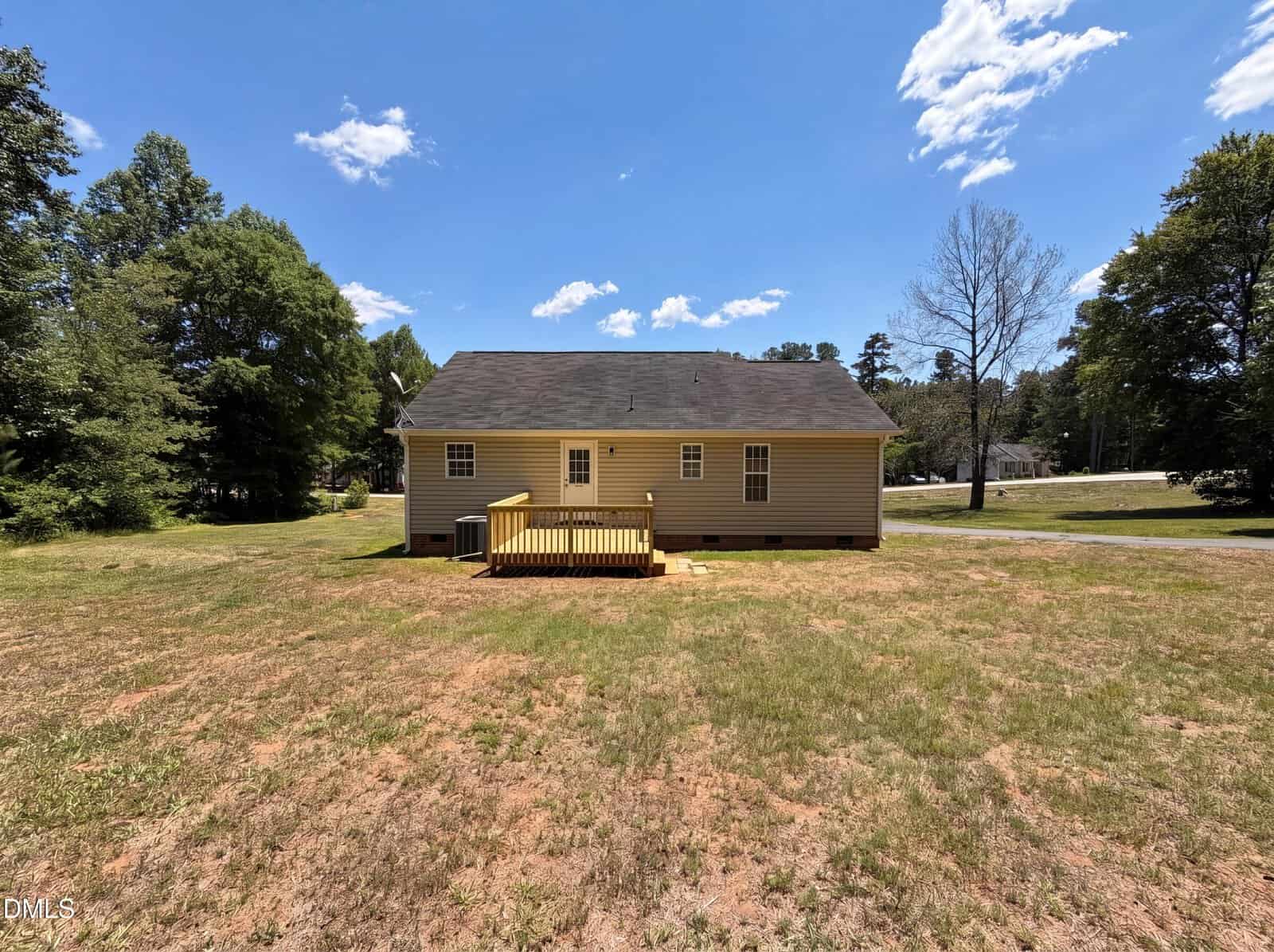 A small yellow house with a black roof and a wooden deck sits on a large, mostly barren yard with sparse grass, surrounded by trees under a bright blue sky with scattered clouds.