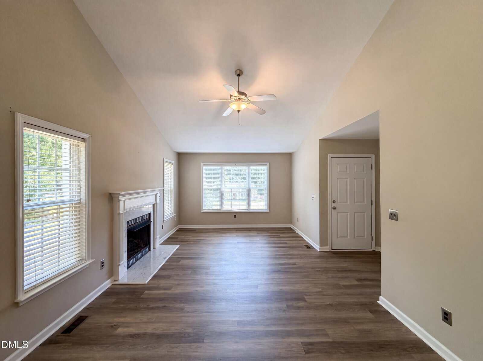 A spacious living room with light wood flooring, beige walls, a white fireplace on the left, ceiling fan above, and large windows at the far end letting in natural light.