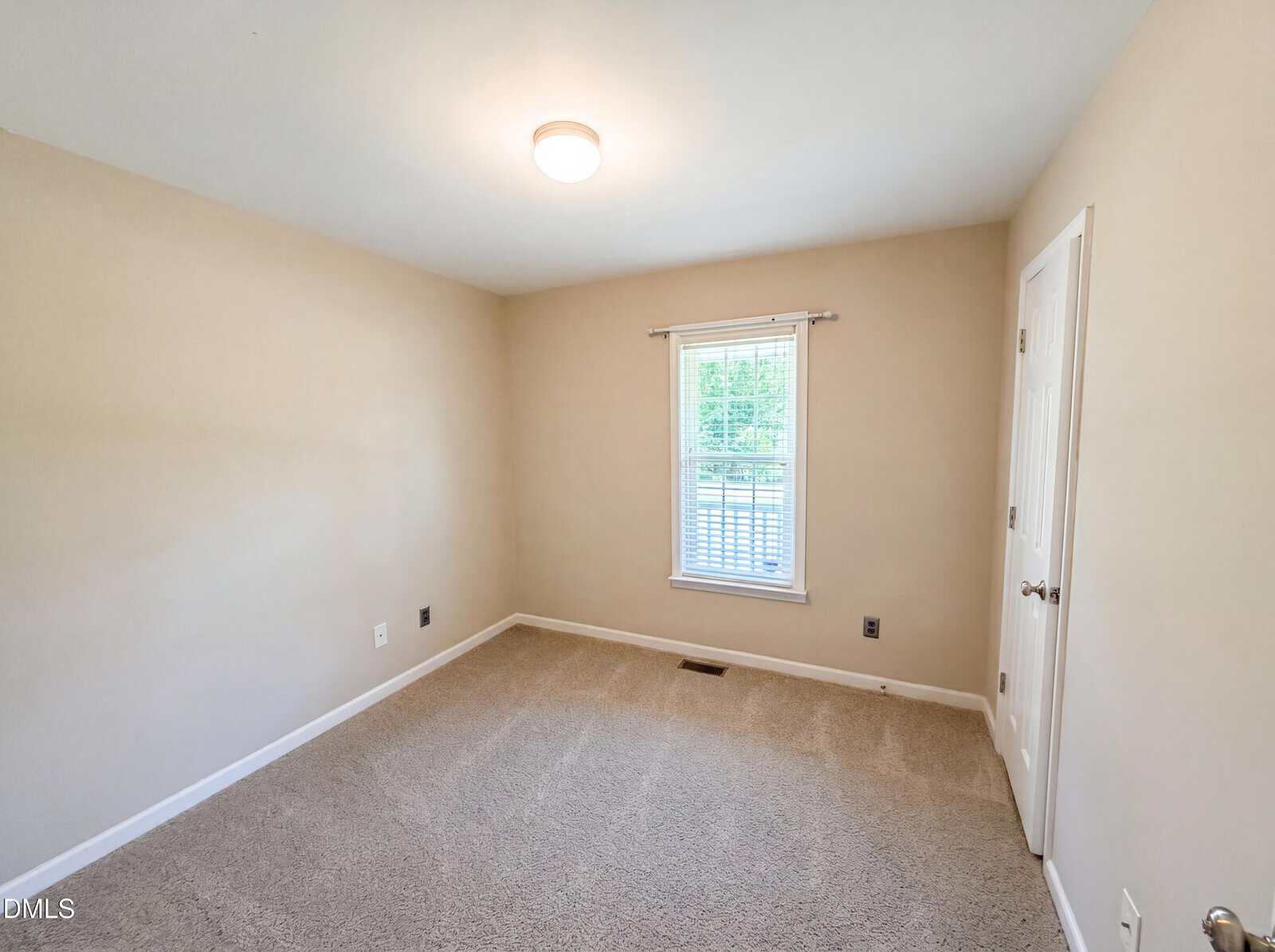 A small, empty room with beige walls, a window with blinds, a closed white door, and beige carpet flooring. A ceiling light is turned on, and sunlight enters through the window.