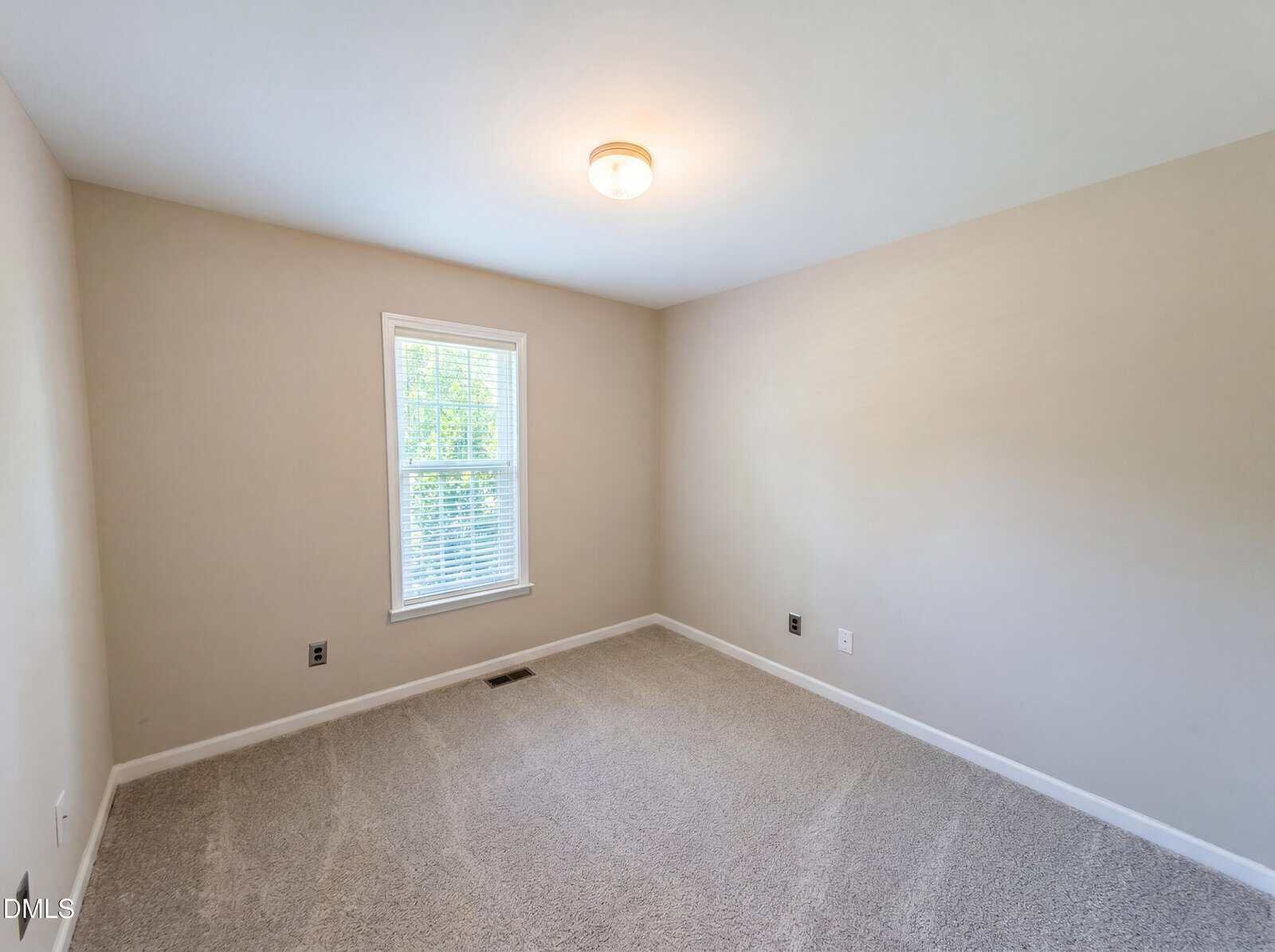 Empty room with beige walls, a single window with white blinds, light gray carpet, white ceiling, and a ceiling light fixture. Natural light enters from the window on the left wall.