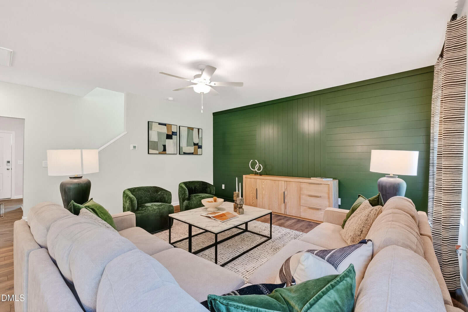 Modern living room with a beige sectional sofa, green accent chairs, a coffee table, and lamps. The back wall is dark green with wood paneling, and a light wood sideboard sits below decor and art.