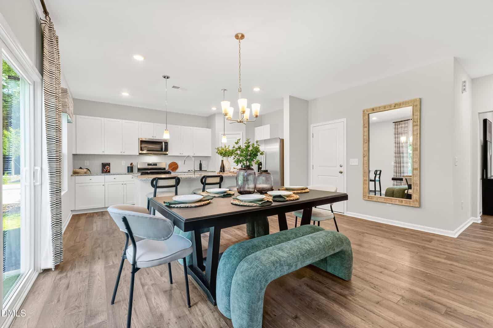 Modern dining area with a black table, four chairs, and a green bench. Kitchen with white cabinets and stainless steel appliances in the background. Large mirror on the wall, wood floors, and natural light from sliding glass doors.