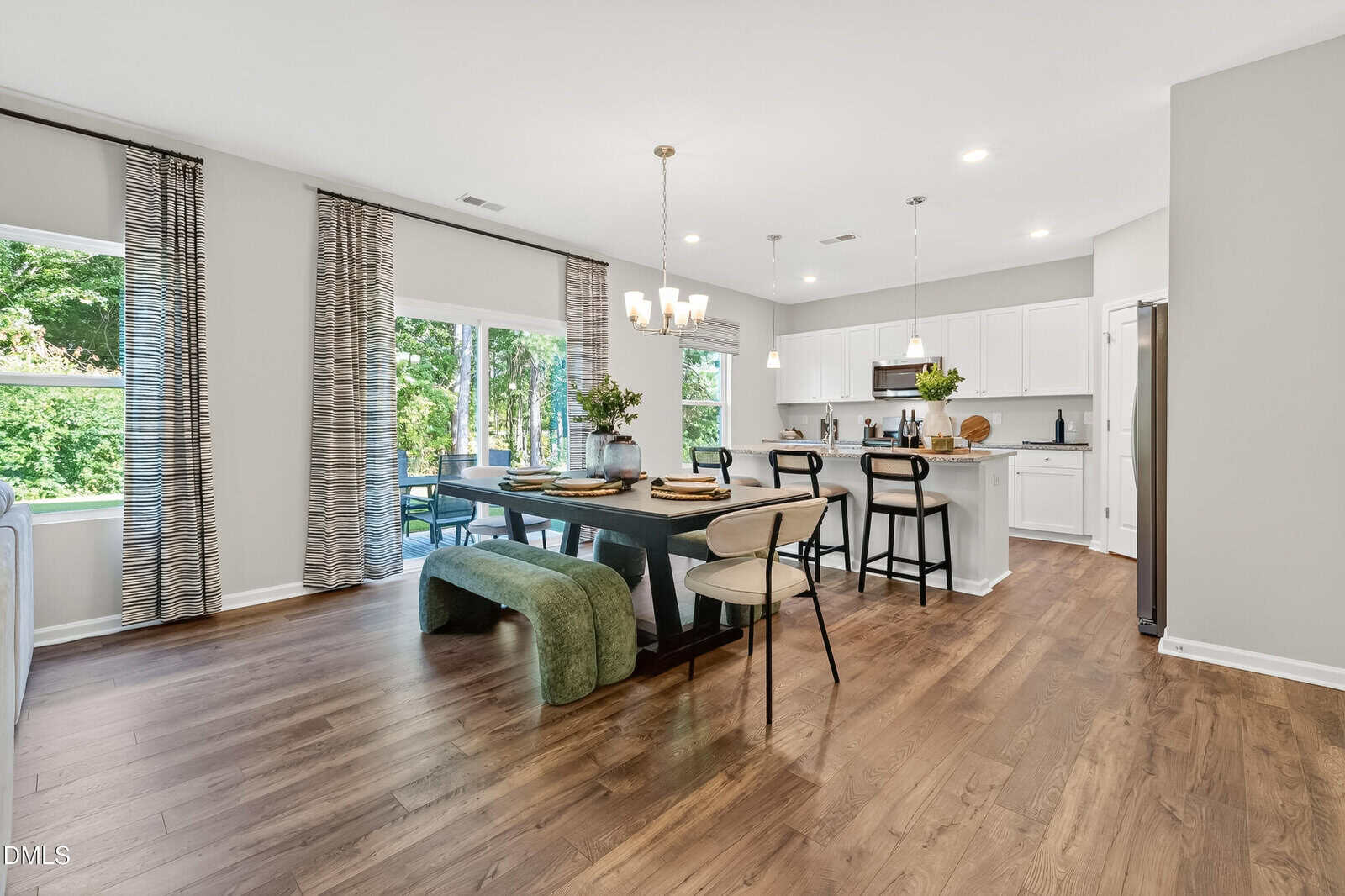 Modern open-plan kitchen and dining area with wooden floors, white cabinets, a large dining table set for four, green upholstered bench, and large windows overlooking greenery. Bright, airy, and welcoming space.