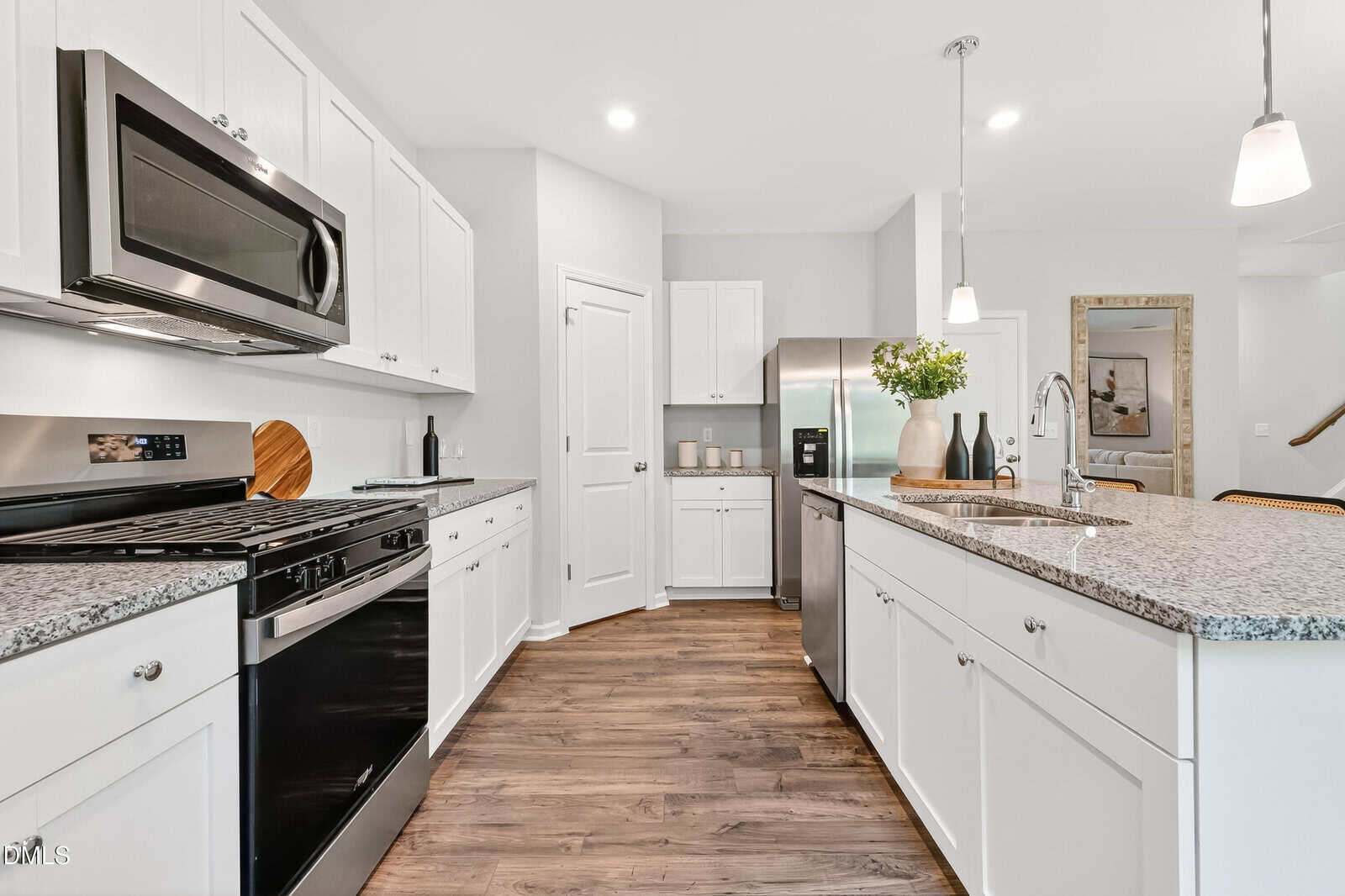 Modern kitchen with white cabinets, stainless steel appliances, granite countertops, a kitchen island with sink, pendant lighting, and wood flooring. A plant sits on the island and a painting hangs on the wall in the background.