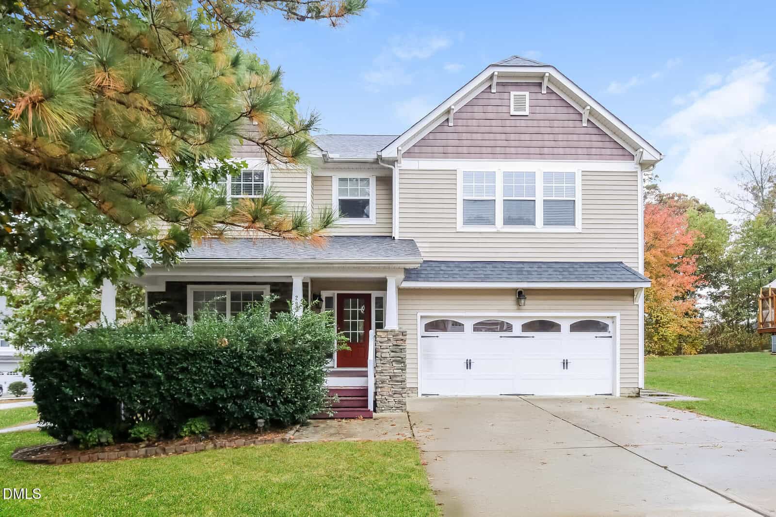Located at 295 Clubhouse Drive in Youngsville, this two-story suburban house features tan siding, white trim, and a double garage. A covered porch with stone columns overlooks green bushes and a well-kept lawn, framed by trees and blue sky.