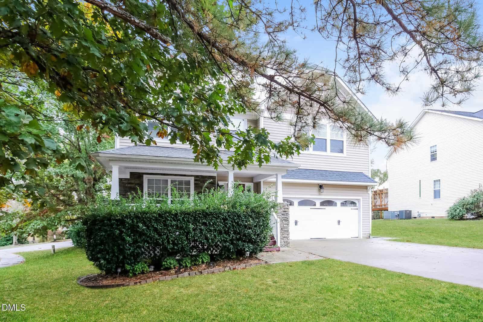 Two-story suburban house with light siding, a covered front porch, and a double garage. Lush green lawn, shrubs, and mature trees frame the driveway and entrance.