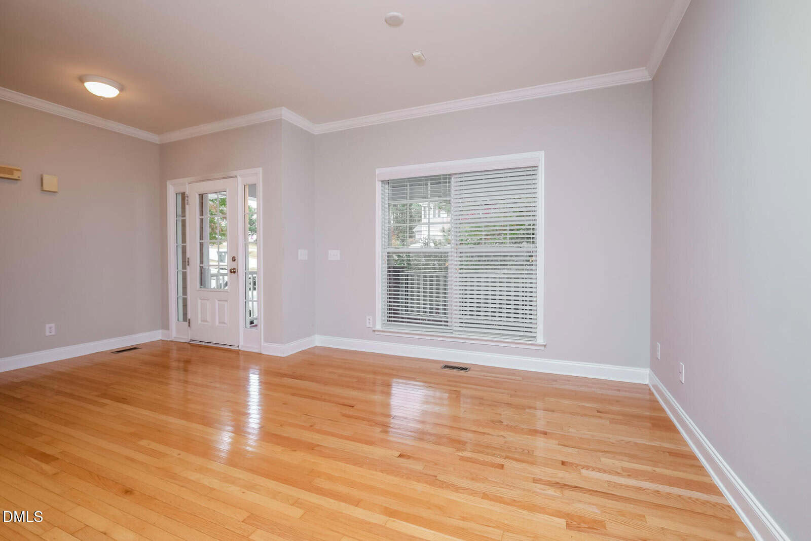 Empty room with light gray walls, hardwood floors, a large window with white blinds, and double French doors. The ceiling has crown molding and a light fixture, and natural light fills the space.