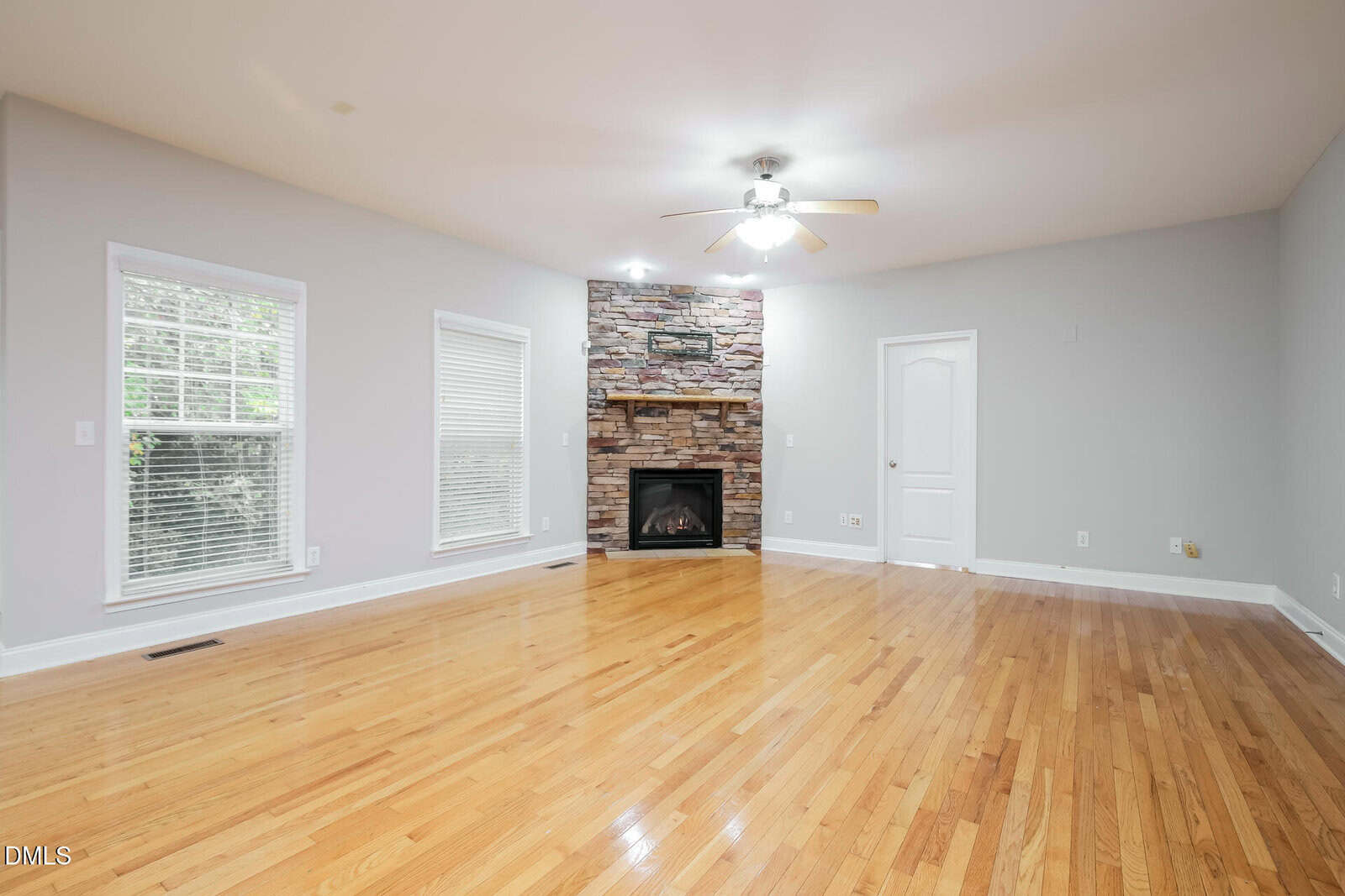 Spacious living room with light hardwood floors, two tall windows, a ceiling fan with light, and a stone fireplace centered on the far wall. Walls are painted light gray and there is a white door to the right of the fireplace.