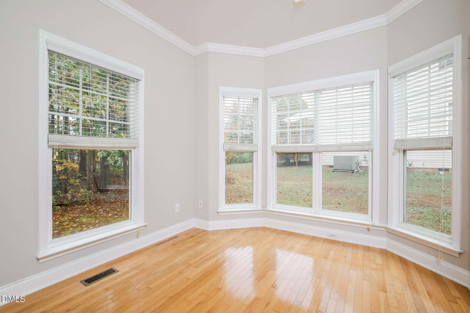 A bright room with light-colored walls and hardwood floors, featuring three large windows with white blinds, offering a view of a green, tree-filled backyard.