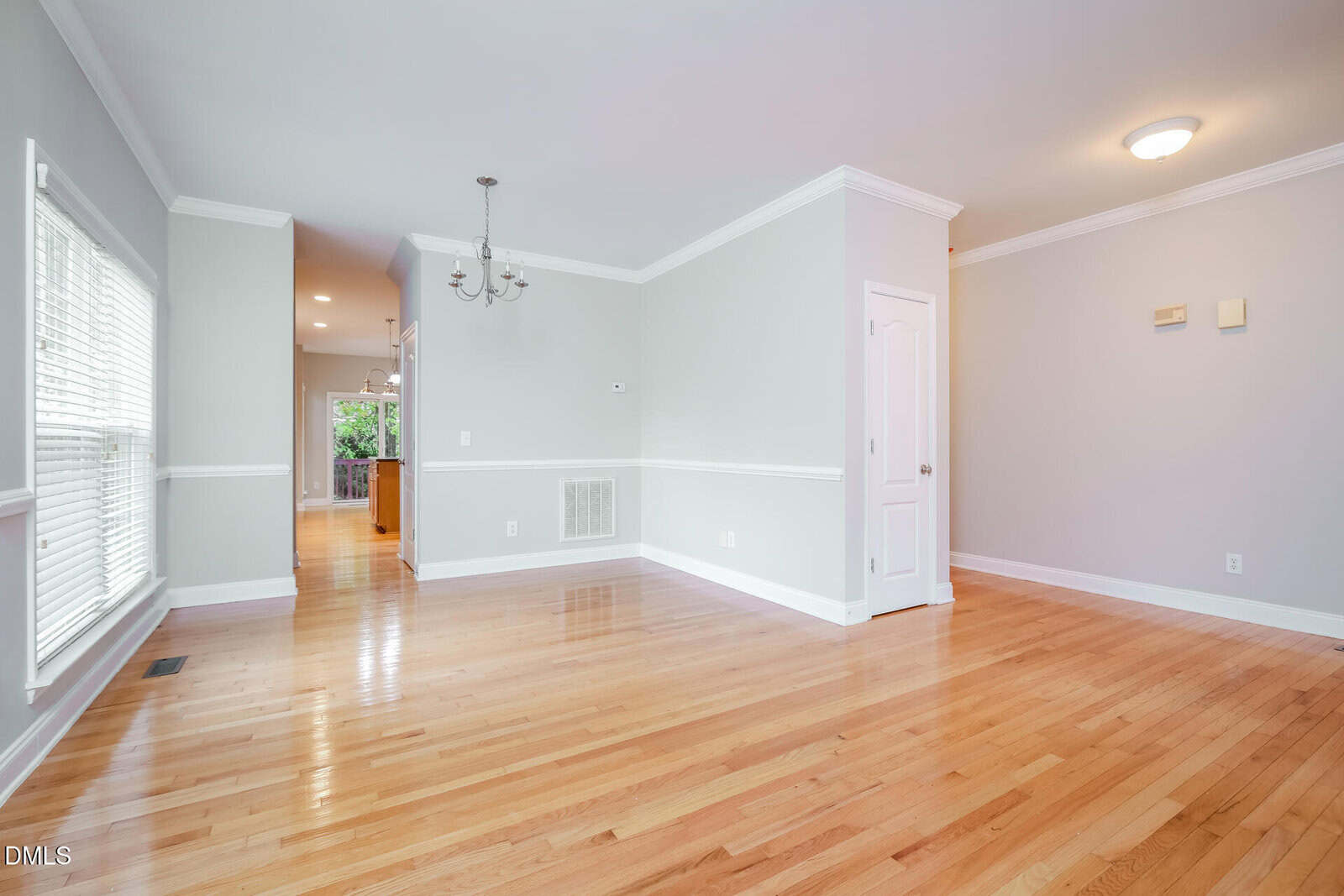 Bright, empty room with light gray walls, white trim, hardwood floor, large windows on the left, and a small chandelier hanging from the ceiling. A hallway leads to another area with visible kitchen cabinets.