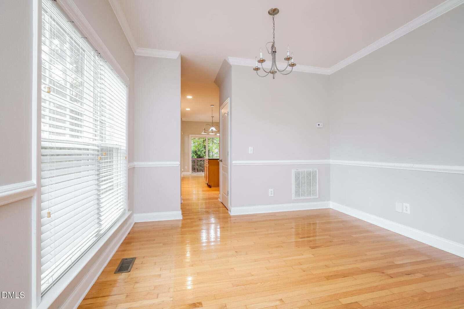Bright dining area with hardwood floors, a small chandelier, and large windows. The space opens to a hallway leading to a kitchen with hanging lights and cabinets visible in the background. Walls are light gray with white trim.