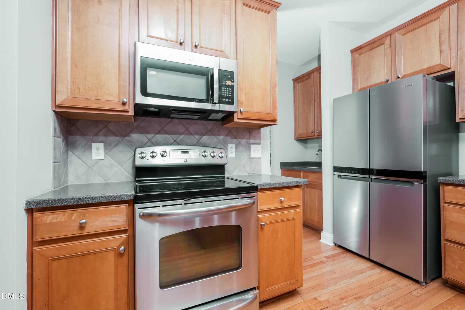 Modern kitchen with stainless steel appliances, including an oven, microwave, and refrigerator. Wood cabinets, dark countertops, and light tiled backsplash on a hardwood floor.
