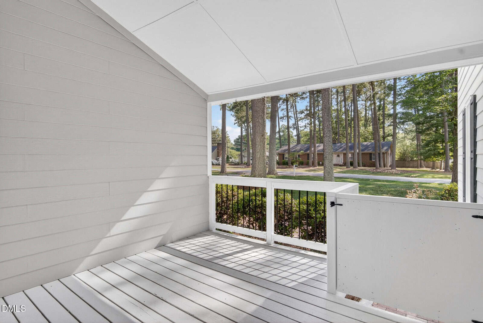 A white, covered front porch with a half gate, white railing, and view of a grassy yard and houses across the street lined with tall trees.
