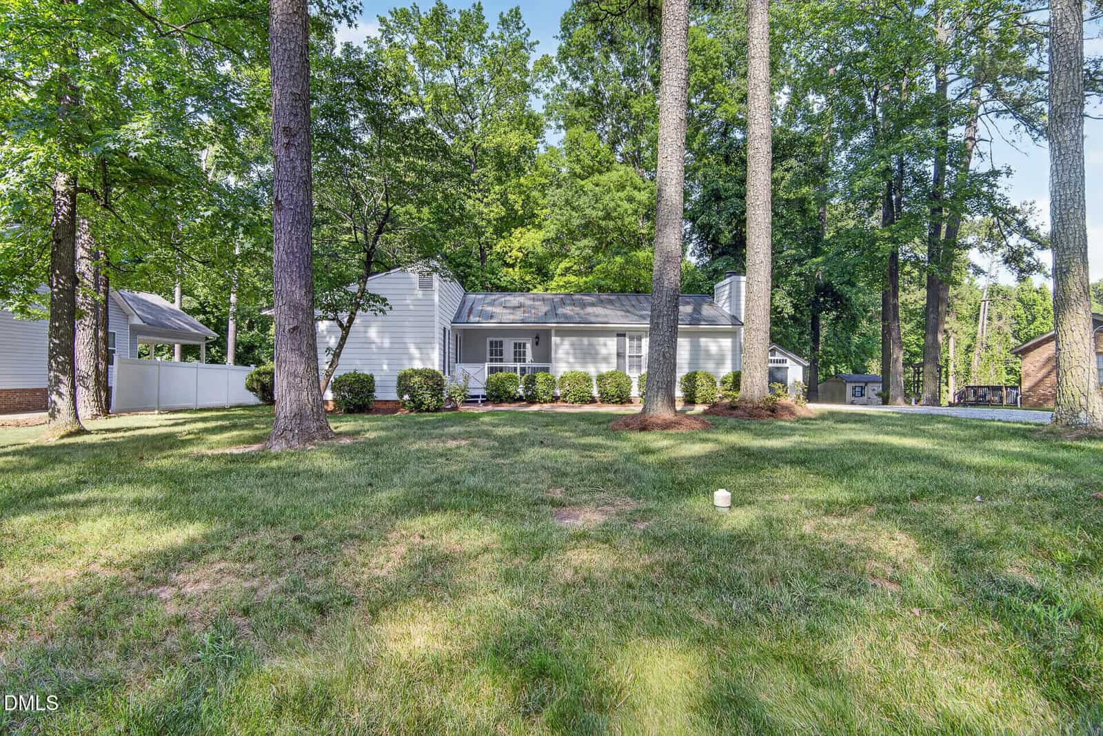 Single-story white house with gray roof, surrounded by tall trees and bushes, sits in a spacious grassy yard on a sunny day. A white fence and driveway are visible on the left side.