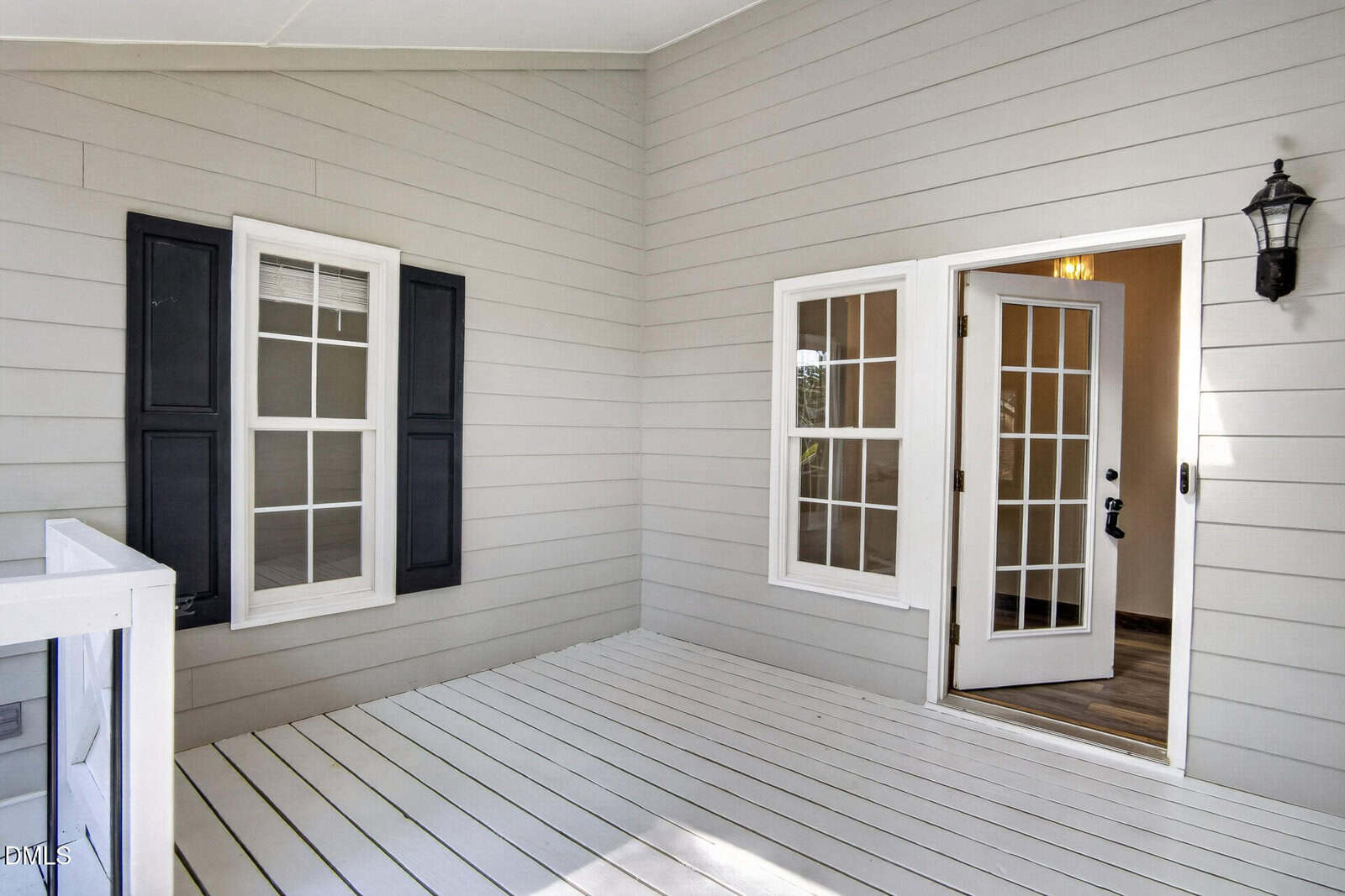 A small porch with white wooden floor, light gray siding, two windows with black shutters, a wall lantern, and open French doors leading inside to a room with wood flooring.