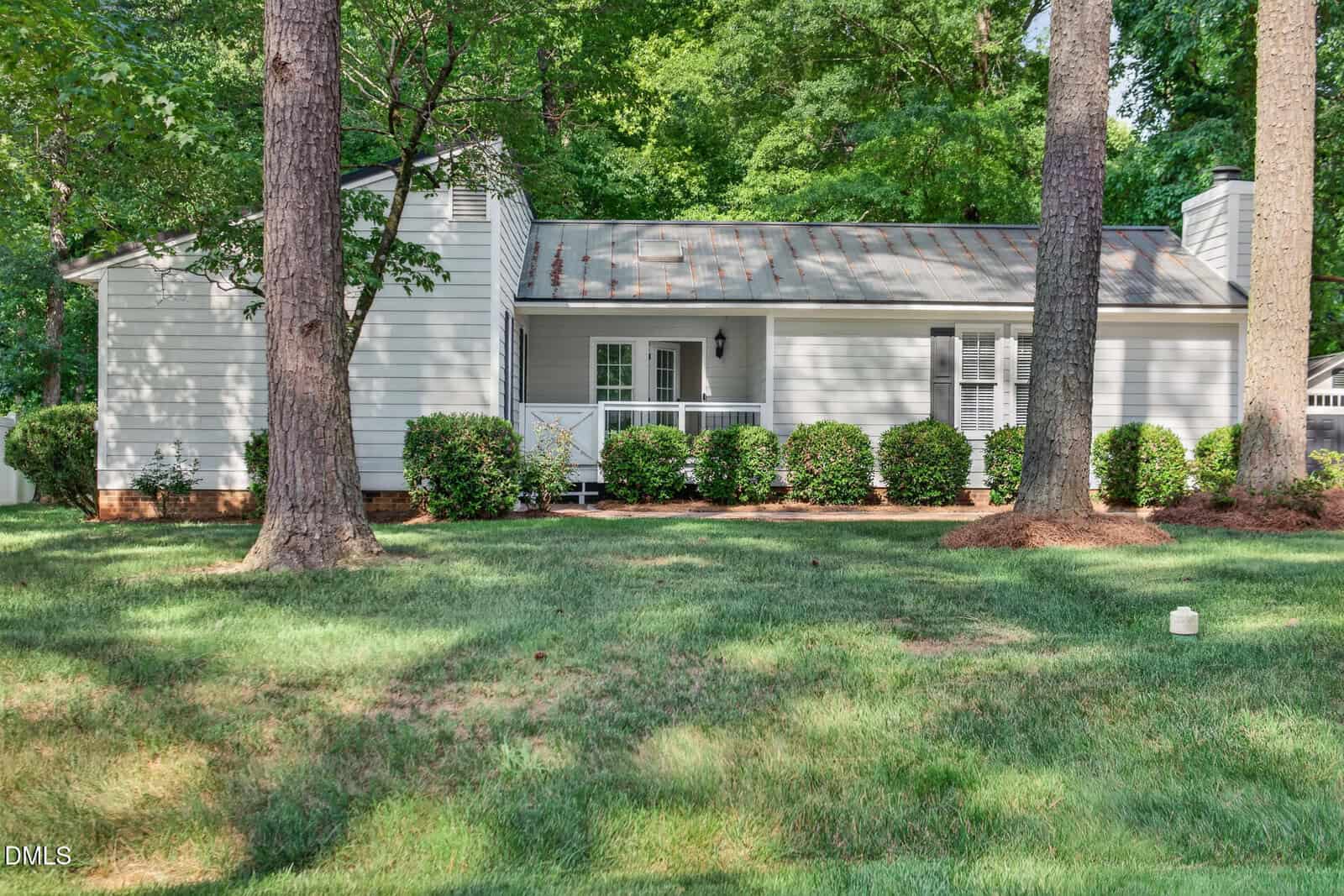 Single-story white house with a metal roof, black shutters, and a small covered porch, surrounded by green grass, shrubs, and tall trees in a shaded yard.