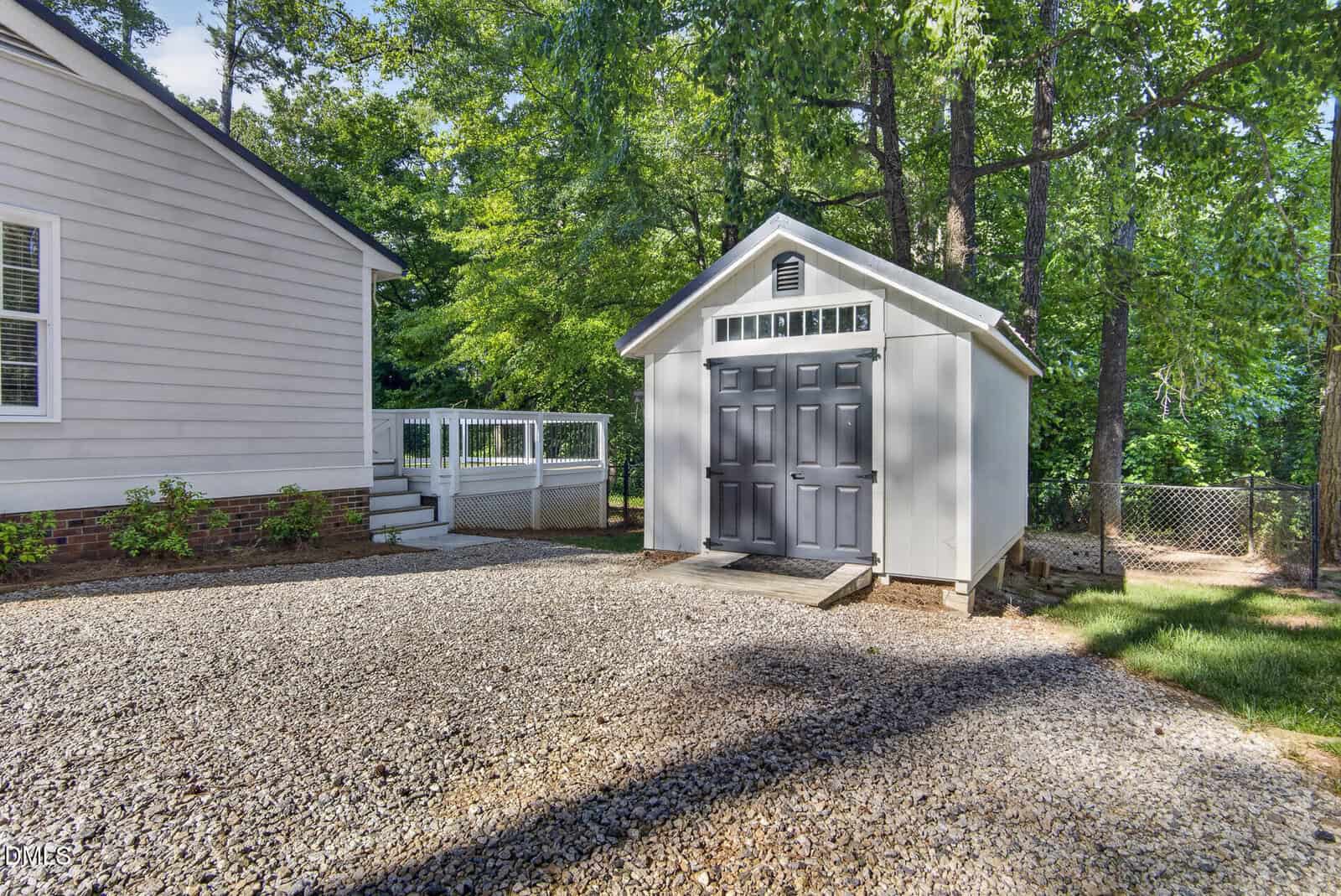 A small white shed with double gray doors sits on a gravel driveway next to a light gray house with white trim, surrounded by trees and greenery.