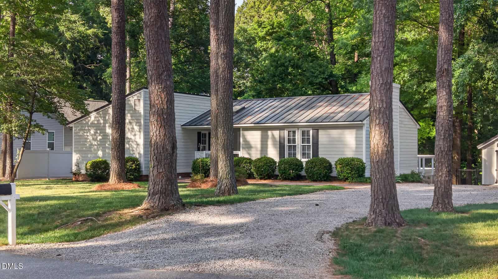 Nestled in Henderson, 165 Dorsey Place is a single-story light gray house with a metal roof, set behind tall trees and lush greenery, with a curved gravel driveway welcoming you to the front.