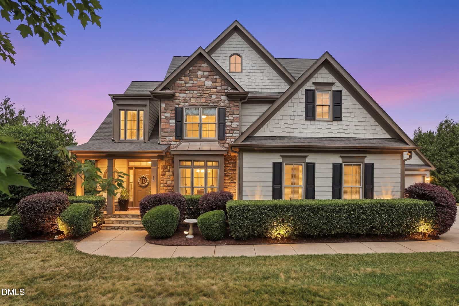 Two-story house with stone and wood siding, multiple gables, shuttered windows, manicured bushes, and evening lighting against a purple sunset sky in Youngsville at 55 Princeton Manor Drive.