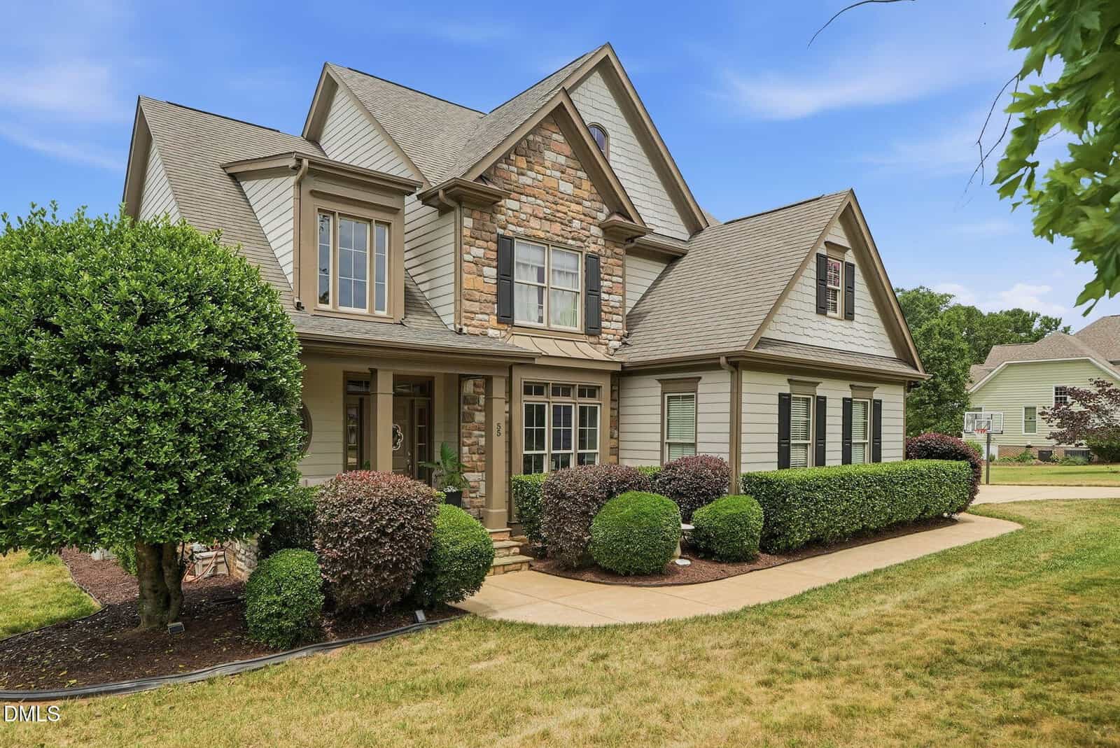 Two-story suburban house with stone and beige siding, gabled roof, front porch, and well-maintained shrubs and lawn under a blue sky.