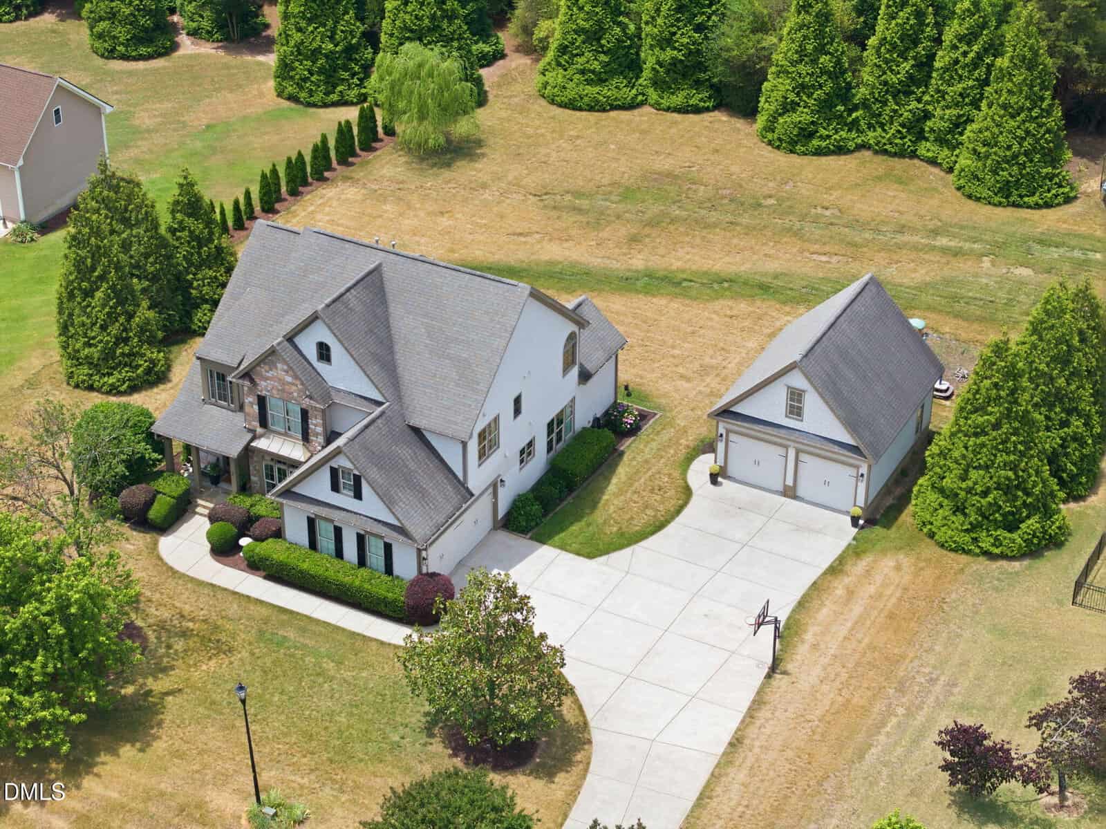 Aerial view of a two-story house with gray roof, detached two-car garage, long driveway, green trees, and neatly trimmed lawn in a suburban neighborhood.