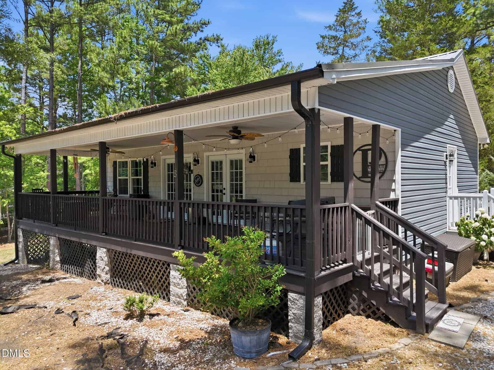 Nestled in Louisburg, 113 Desoto Drive is a cozy gray house with white trim, a spacious covered porch with ceiling fans, dark railings, and steps leading to a gravel yard. Surrounded by trees and greenery under a bright blue sky.