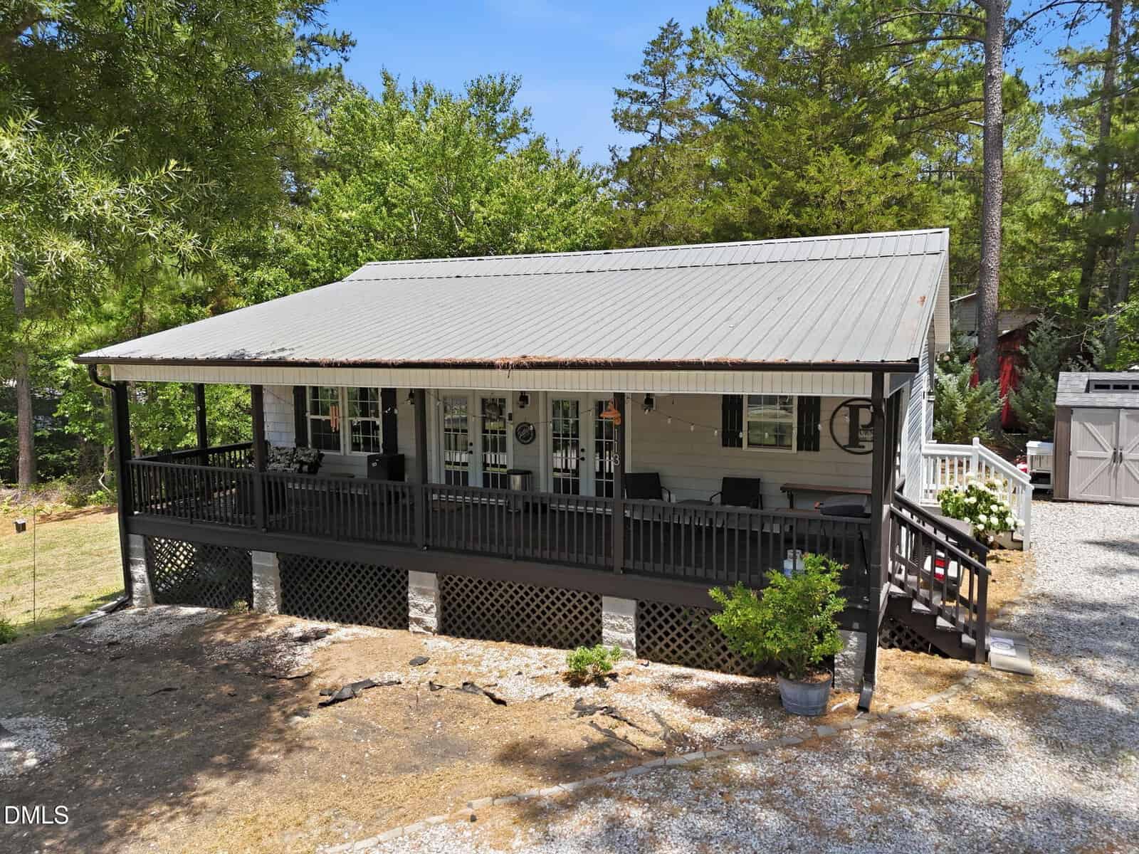 A single-story house with a large covered front porch, black railing, and steps leading down to a gravel driveway, surrounded by trees and greenery.
