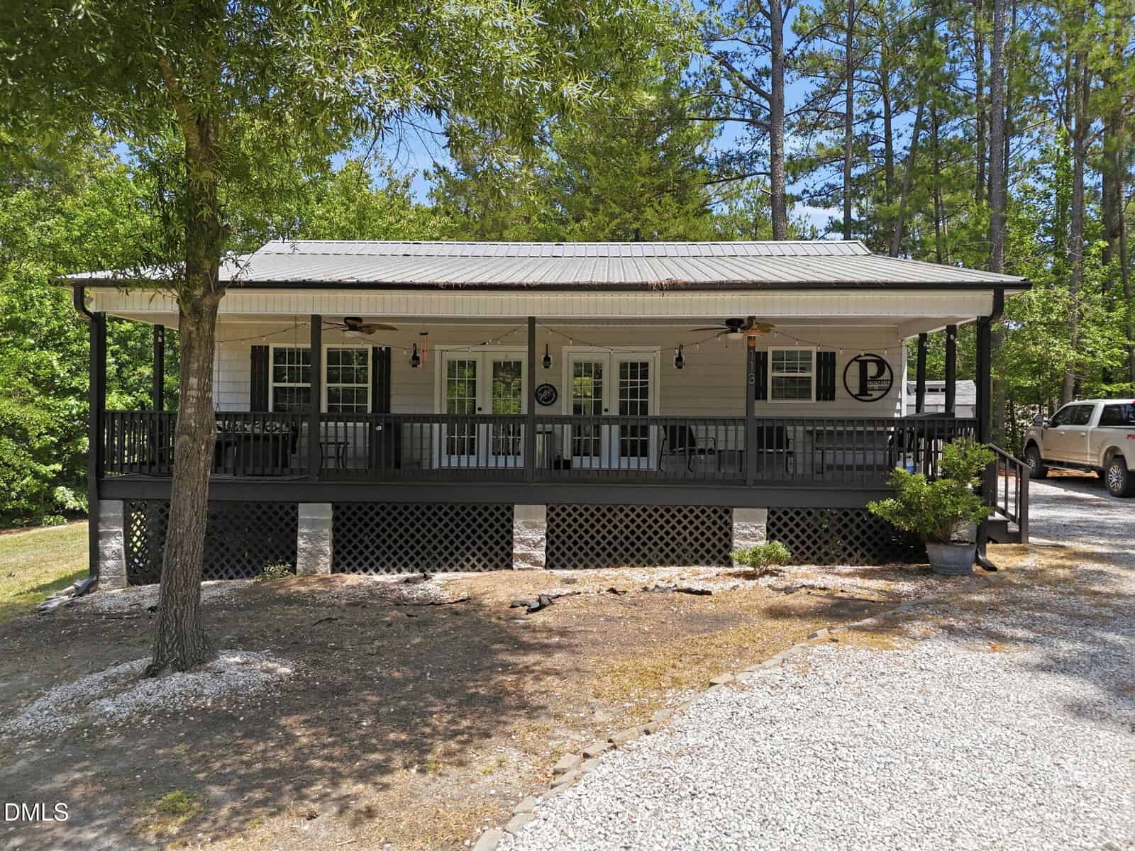 A single-story house with a large covered front porch, dark railings, and outdoor ceiling fans, surrounded by trees. A gravel driveway and parked vehicles are visible beside the house.