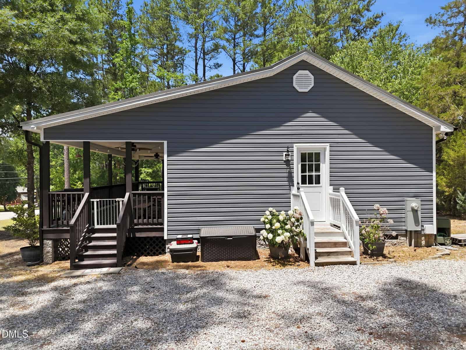 A single-story gray house with white trim, a small porch with stairs on the right, a covered side porch on the left, and white gravel driveway, surrounded by trees and greenery.