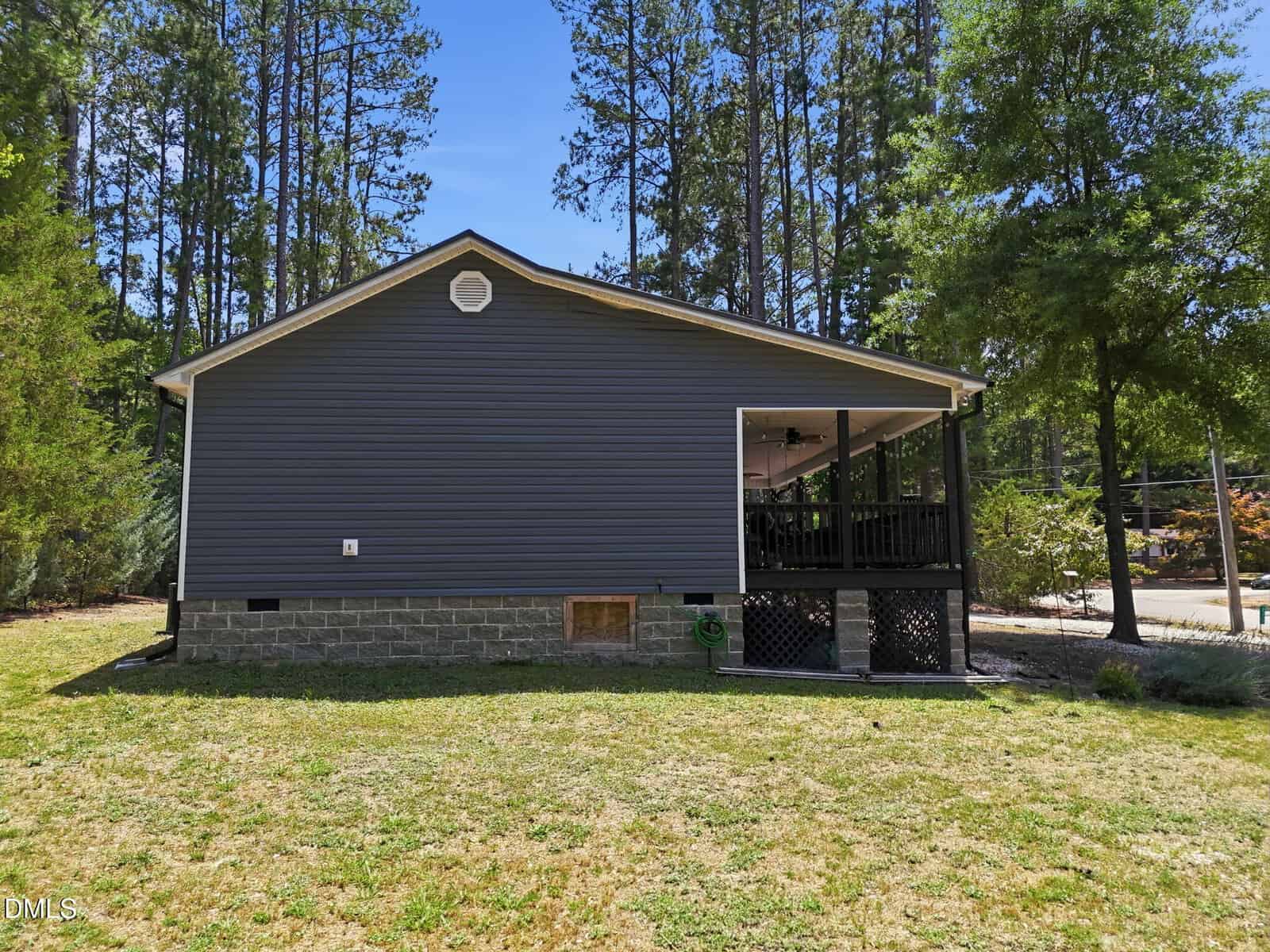 A side view of a gray house with a screened porch, surrounded by grass, trees, and a forested area in the background under a clear blue sky.
