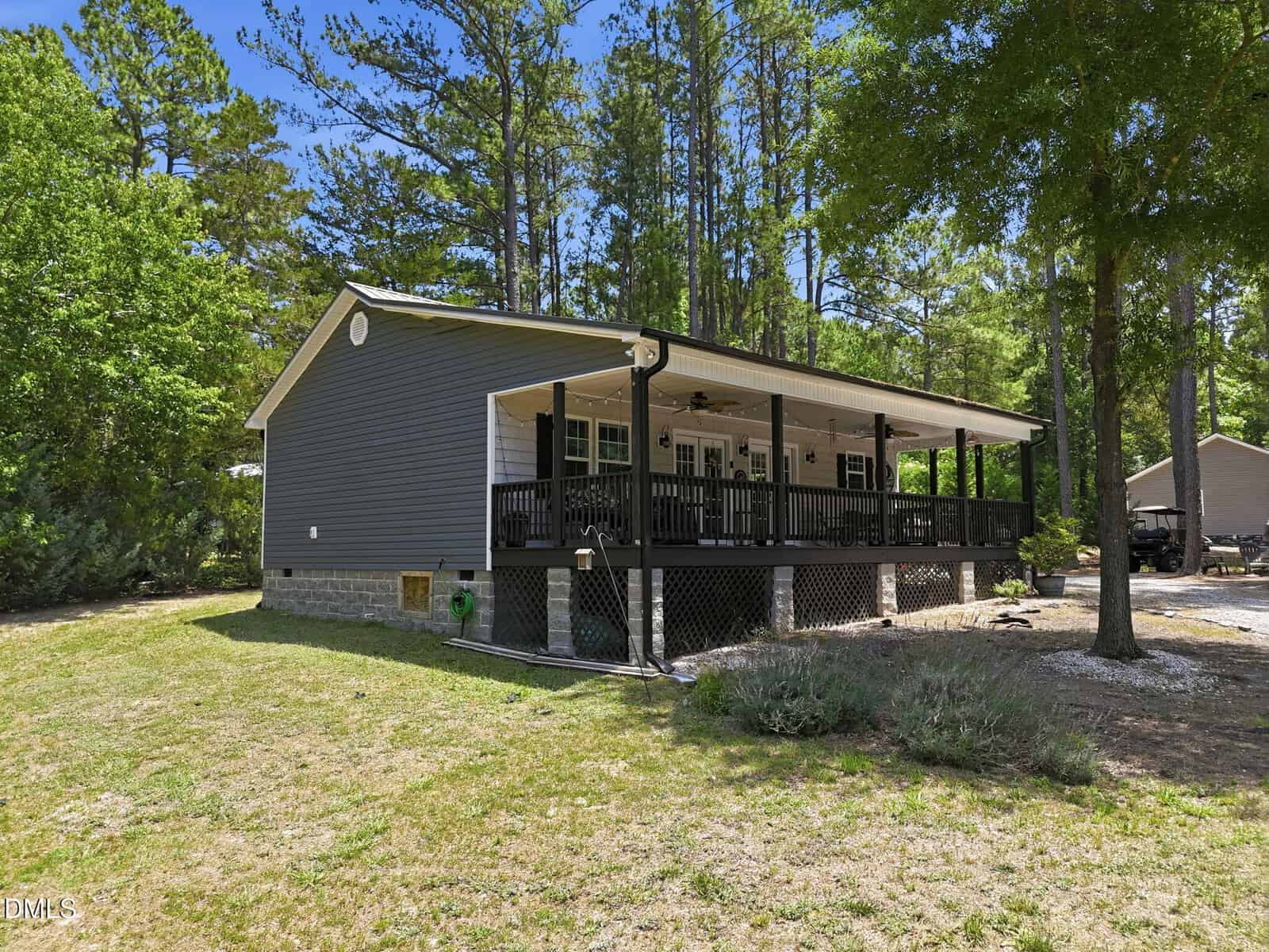 A gray single-story house with a large covered front porch, black railing, and ceiling fans, set in a grassy, wooded area with trees and blue sky in the background.