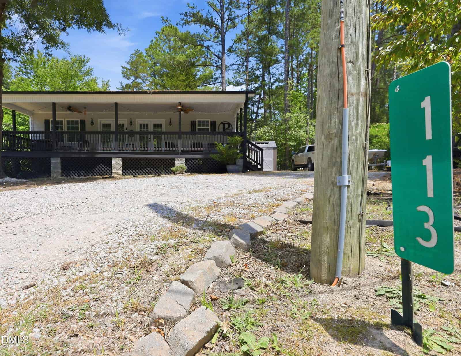 A gravel driveway leads to a house with a large covered porch and ceiling fans. A green sign with the numbers “113” is posted by a utility pole in the foreground. Tall trees surround the property.