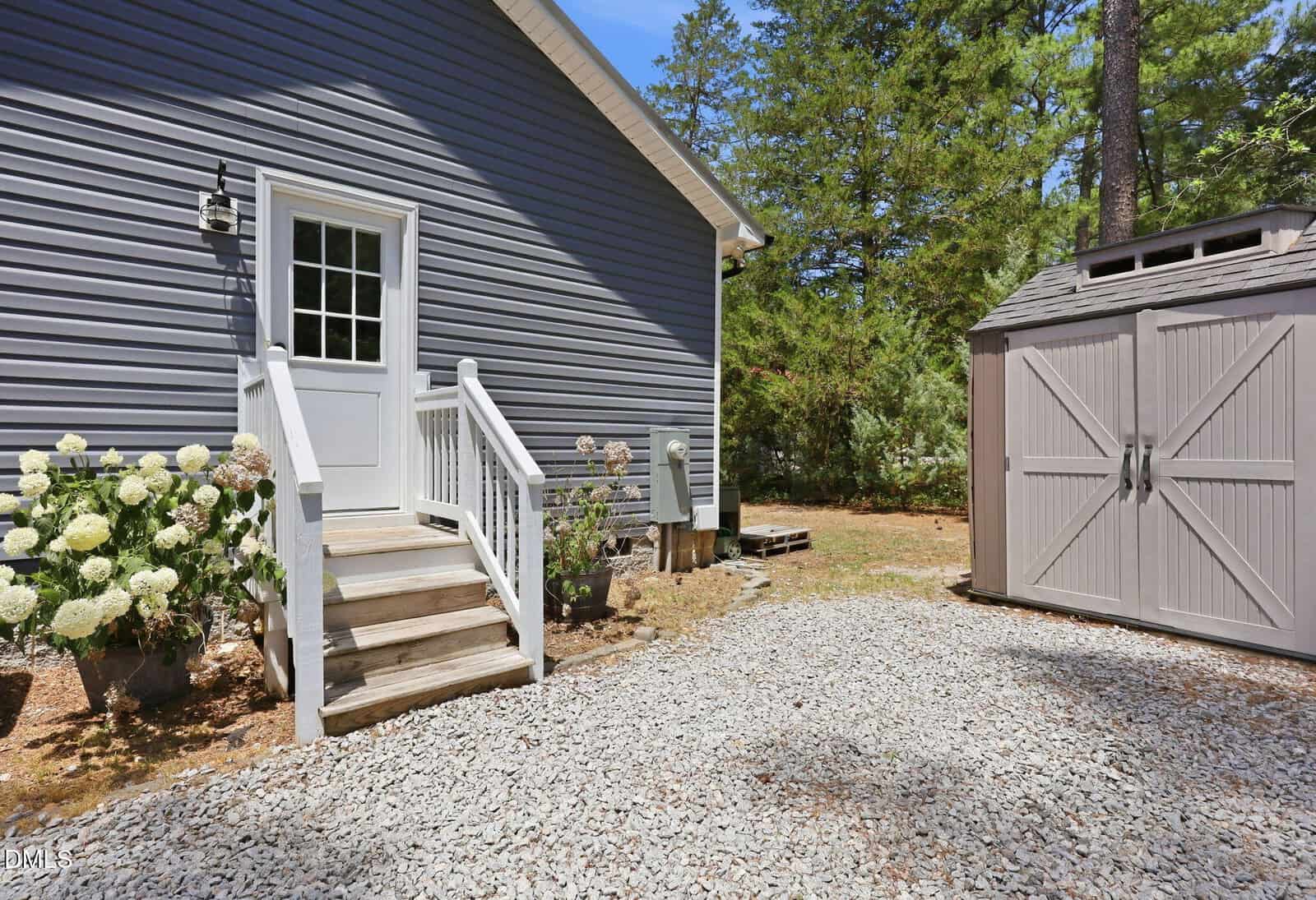 A small set of white stairs leads to a door on a gray house with white trim. There are potted flowers nearby, a gravel path, and a matching gray storage shed in a yard with trees in the background.