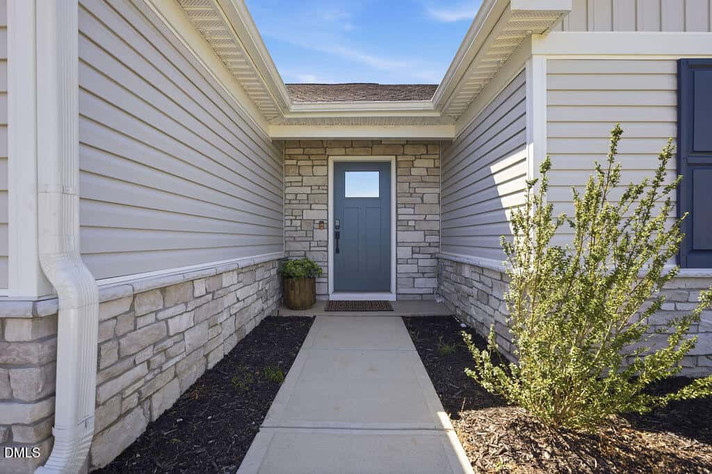 A concrete walkway leads to a blue front door set in a stone facade, flanked by beige siding. A potted plant sits to the left of the door, and a green shrub is on the right side of the path.
