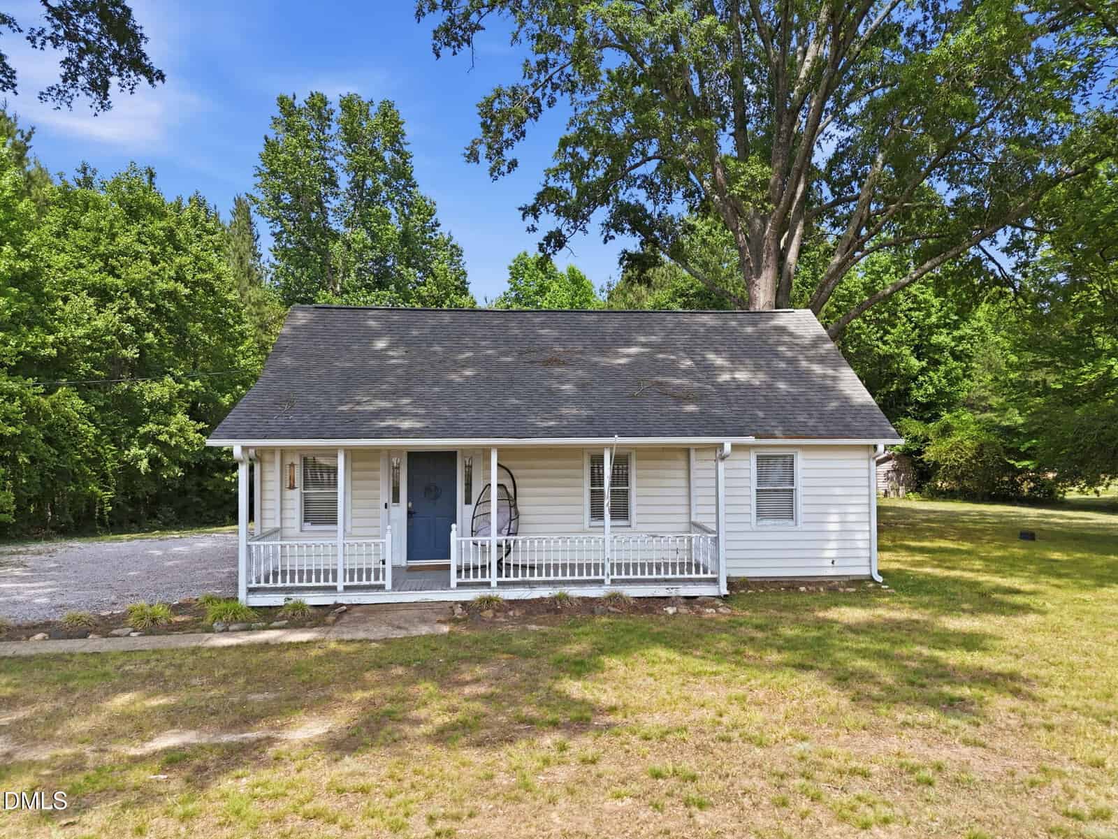A small white house with a gray roof and a covered front porch sits on a grassy lot surrounded by trees under a clear blue sky.