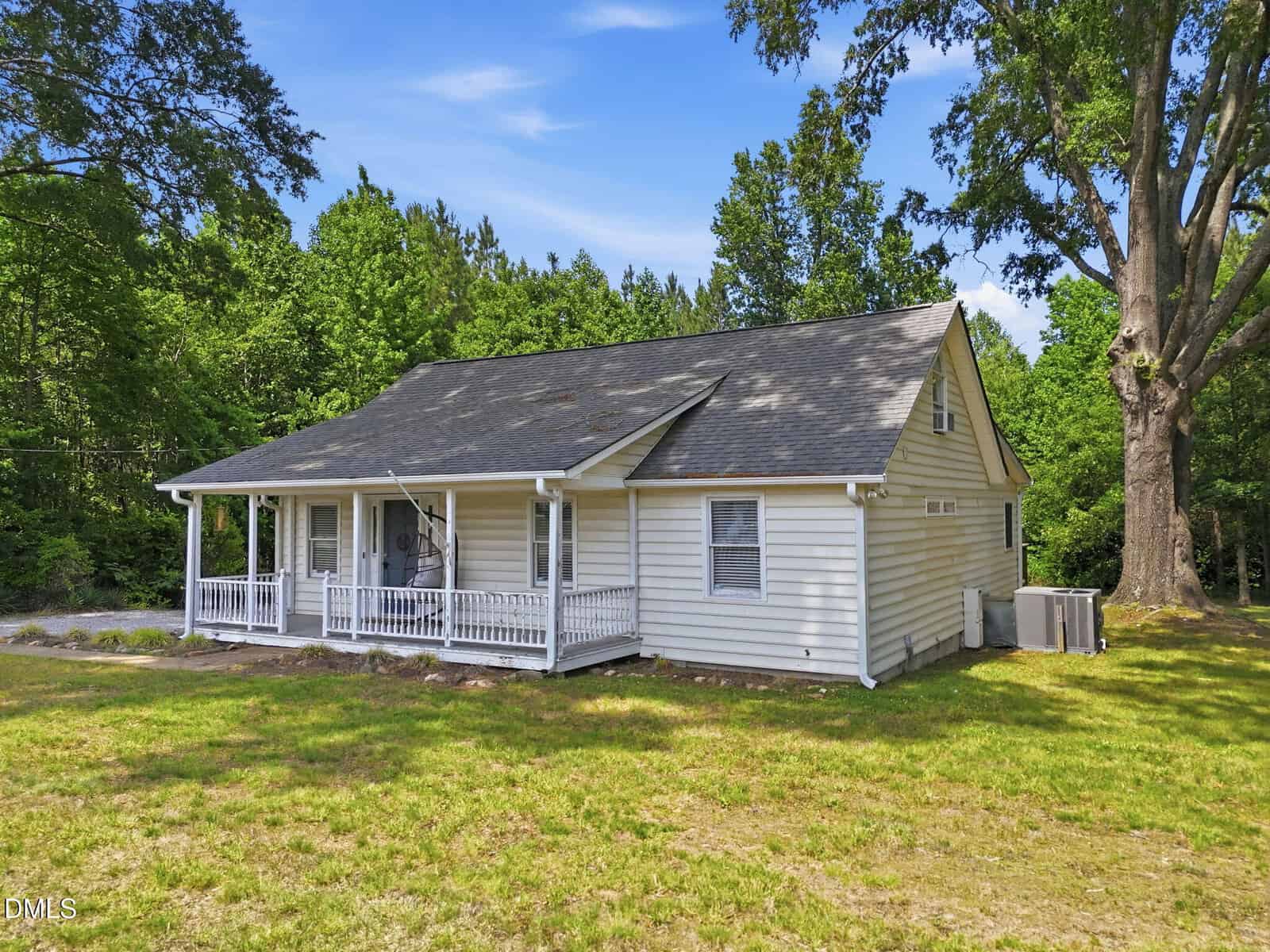 Charming single-story white house with a gray roof at 2568 NC 96 S, Franklinton. Features a front porch with railing, set on a grassy yard with large trees and lush greenery beneath a clear blue sky.