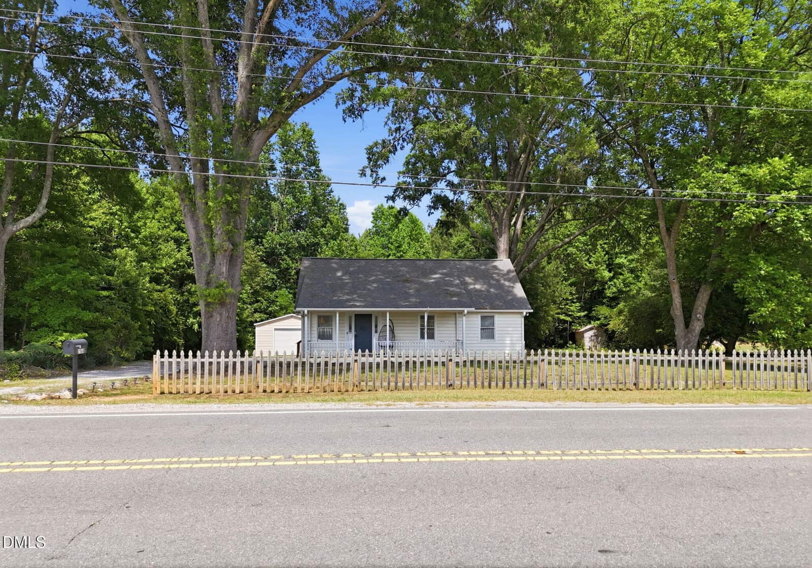 Small white house with a gray roof and front porch, surrounded by tall trees and a wooden picket fence, sits behind a two-lane road on a sunny day. A mailbox and detached garage are visible on the left.
