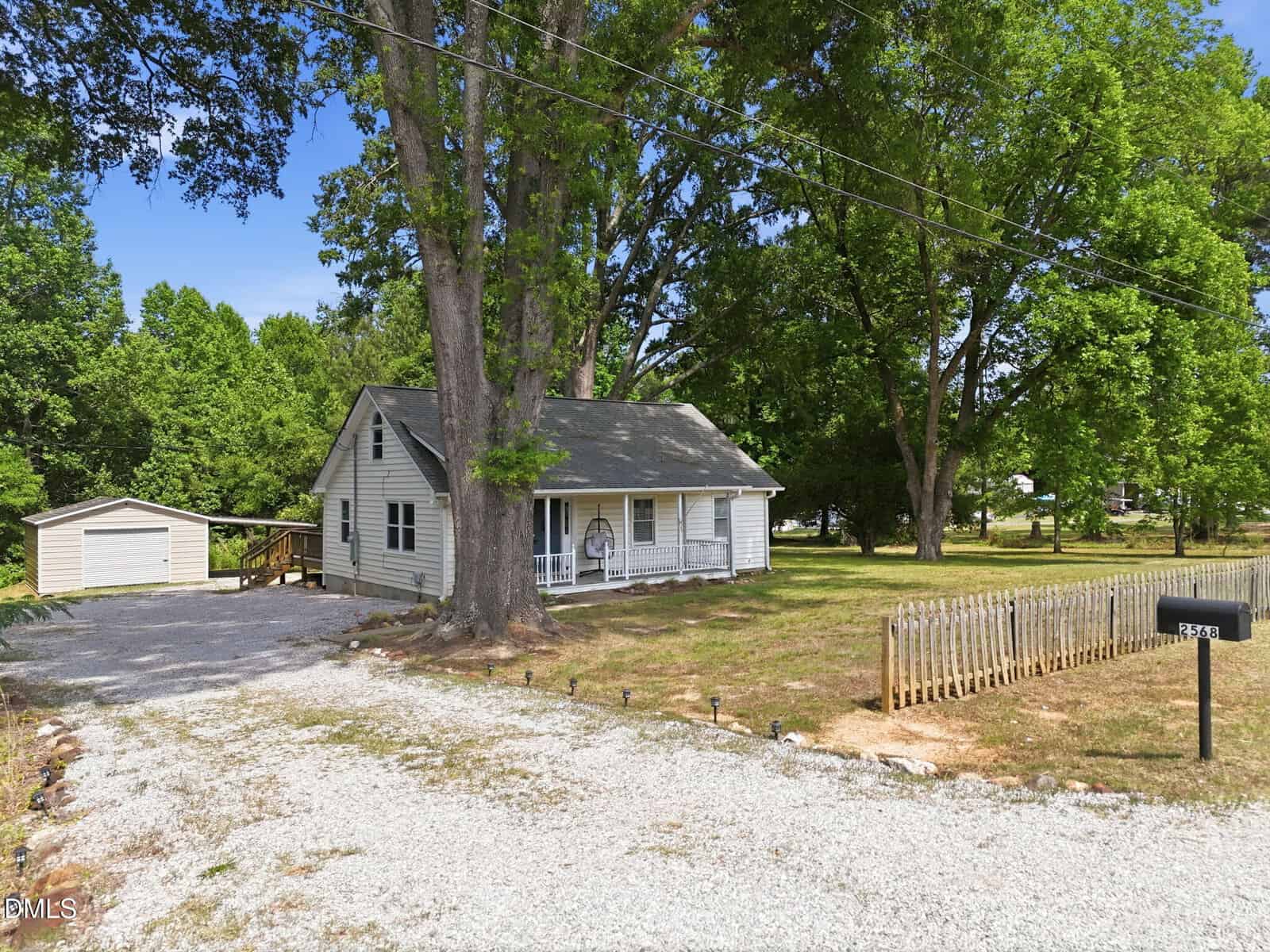 A small gray house with a covered front porch sits among tall trees, next to a gravel driveway leading to a detached garage and a black mailbox. The yard is mostly grass with patches of bare ground and a small wooden fence.